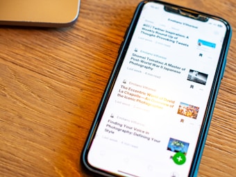 A smartphone displaying a list of articles with their titles, authors, and reading times is placed on a wooden surface. The phone screen is illuminated, and a part of a laptop is visible in the background.