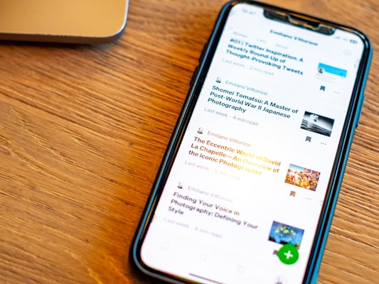 A Smartphone Displaying A List Of Articles With Their Titles, Authors, And Reading Times Is Placed On A Wooden Surface. The Phone Screen Is Illuminated, And A Part Of A Laptop Is Visible In The Background.