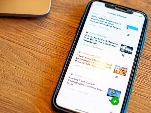 A smartphone displaying a list of articles with their titles, authors, and reading times is placed on a wooden surface. The phone screen is illuminated, and a part of a laptop is visible in the background.