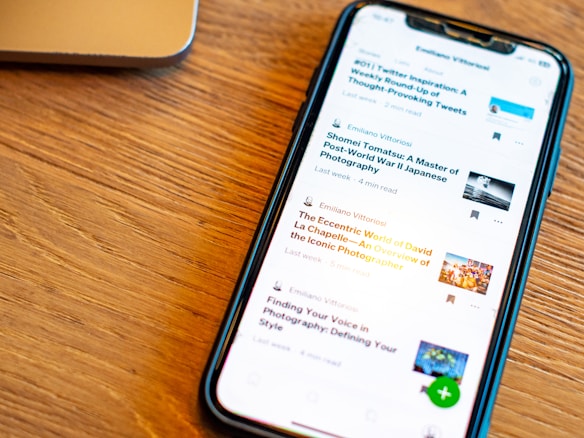 A smartphone displaying a list of articles with their titles, authors, and reading times is placed on a wooden surface. The phone screen is illuminated, and a part of a laptop is visible in the background.