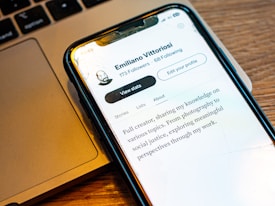 A smartphone displays a profile on a social media application. The profile includes the name Emiliano Vittoriosi, the follower count, and a profile summary stating interests in photography and social justice. The phone is placed on a MacBook laptop, with part of the keyboard visible.