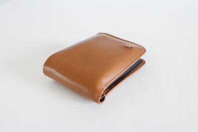 A brown leather wallet with a simple, sleek design is positioned on a plain white surface. The wallet is closed, and a small metallic logo accent is visible on the upper flap.