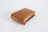 A brown leather wallet with a simple, sleek design is positioned on a plain white surface. The wallet is closed, and a small metallic logo accent is visible on the upper flap.