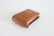 A brown leather wallet with a simple, sleek design is positioned on a plain white surface. The wallet is closed, and a small metallic logo accent is visible on the upper flap.