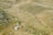 A couple walks through a grassy canyon landscape, with sparse vegetation scattered across rolling hills. The scene appears tranquil, with the earthy tones of the grass blending into the surroundings. The couple, dressed formally, suggests a special occasion or moment.