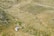 A couple walks through a grassy canyon landscape, with sparse vegetation scattered across rolling hills. The scene appears tranquil, with the earthy tones of the grass blending into the surroundings. The couple, dressed formally, suggests a special occasion or moment.