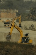 A construction site with two large yellow excavators working amidst sand dunes. A person is visible inside the cab of the closer excavator, which has its arm extended into a trench. In the background, there is a brick building with a flat roof surrounded by vegetation, including trees and tall grass.