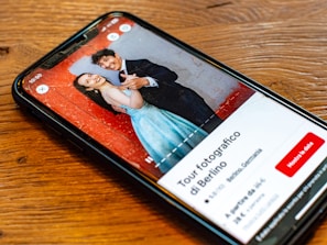 A smartphone lying on a wooden table displaying an image of a smiling couple. The man and woman are dressed formally; he is wearing a black suit, and she is in a blue dress. The background in the photo is a textured red wall. The screen also shows text in Italian, along with buttons and a rating for a photography tour in Berlin.