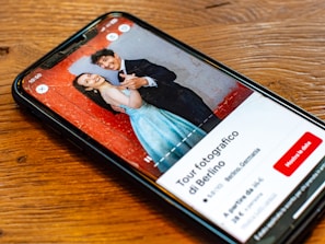 A smartphone lying on a wooden table displaying an image of a smiling couple. The man and woman are dressed formally; he is wearing a black suit, and she is in a blue dress. The background in the photo is a textured red wall. The screen also shows text in Italian, along with buttons and a rating for a photography tour in Berlin.