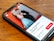A smartphone lying on a wooden table displaying an image of a smiling couple. The man and woman are dressed formally; he is wearing a black suit, and she is in a blue dress. The background in the photo is a textured red wall. The screen also shows text in Italian, along with buttons and a rating for a photography tour in Berlin.