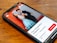 A smartphone lying on a wooden table displaying an image of a smiling couple. The man and woman are dressed formally; he is wearing a black suit, and she is in a blue dress. The background in the photo is a textured red wall. The screen also shows text in Italian, along with buttons and a rating for a photography tour in Berlin.