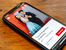 A smartphone lying on a wooden table displaying an image of a smiling couple. The man and woman are dressed formally; he is wearing a black suit, and she is in a blue dress. The background in the photo is a textured red wall. The screen also shows text in Italian, along with buttons and a rating for a photography tour in Berlin.