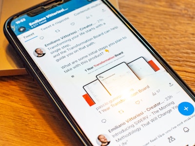 A smartphone displaying a social media post featuring a text discussing transformation and steps to take with a product. The phone is placed on a wooden desk, and there is a partial view of a laptop nearby.