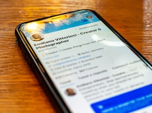 A smartphone lays on a wooden table displaying a social media profile. The screen shows a profile photo, username, and text with details about a creator and photographer. Reflections of overhead lights are visible on the screen.