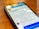 A smartphone lays on a wooden table displaying a social media profile. The screen shows a profile photo, username, and text with details about a creator and photographer. Reflections of overhead lights are visible on the screen.