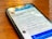 A smartphone lays on a wooden table displaying a social media profile. The screen shows a profile photo, username, and text with details about a creator and photographer. Reflections of overhead lights are visible on the screen.