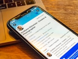 A smartphone displaying a social media profile is placed on a wooden table next to a laptop. The profile includes a picture, name, and details about a creator and photographer. The blue and white interface is visible on the phone screen.