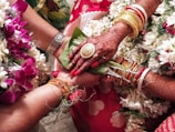 A close-up view of a wedding ritual where two hands are joined together. The bride's hand is adorned with intricate henna designs, gold bracelets, and rings. She holds betel leaves. There are flower garlands in the background, and the atmosphere is colorful and festive.