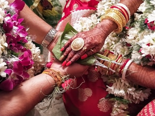 A close-up view of a wedding ritual where two hands are joined together. The bride's hand is adorned with intricate henna designs, gold bracelets, and rings. She holds betel leaves. There are flower garlands in the background, and the atmosphere is colorful and festive.