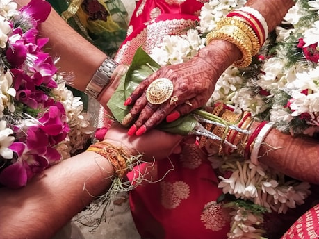 A close-up view of a wedding ritual where two hands are joined together. The bride's hand is adorned with intricate henna designs, gold bracelets, and rings. She holds betel leaves. There are flower garlands in the background, and the atmosphere is colorful and festive.