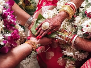 A close-up view of a wedding ritual where two hands are joined together. The bride's hand is adorned with intricate henna designs, gold bracelets, and rings. She holds betel leaves. There are flower garlands in the background, and the atmosphere is colorful and festive.