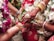 A close-up view of a wedding ritual where two hands are joined together. The bride's hand is adorned with intricate henna designs, gold bracelets, and rings. She holds betel leaves. There are flower garlands in the background, and the atmosphere is colorful and festive.