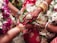 A close-up view of a wedding ritual where two hands are joined together. The bride's hand is adorned with intricate henna designs, gold bracelets, and rings. She holds betel leaves. There are flower garlands in the background, and the atmosphere is colorful and festive.