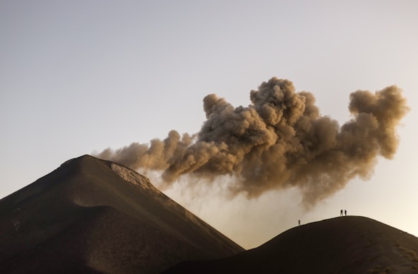 A volcanic landscape with dark, rugged slopes emitting thick clouds of ash into the sky. A couple of silhouetted figures stand on the ridge, observing the scene from a distance.