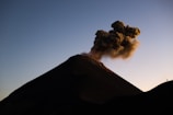 A dramatic aerial shot of an active volcano with smoke rising against a sunset sky.