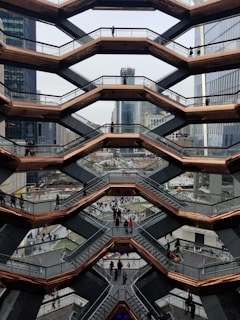 An architectural structure with a honeycomb design features multiple levels of staircases, lined by glass railings and bronze-colored frames. People are walking on the different levels, and the background showcases urban skyscrapers and city buildings.