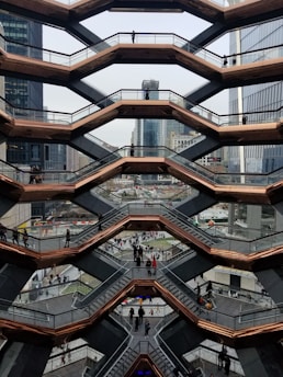 An architectural structure with a honeycomb design features multiple levels of staircases, lined by glass railings and bronze-colored frames. People are walking on the different levels, and the background showcases urban skyscrapers and city buildings.