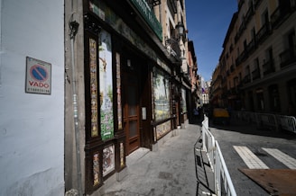 A narrow street lined with historic buildings with ornate facades. A colorful tile mural decorates one wall, and there is a 'no parking' sign nearby. The street is bordered by metal barriers, and some pedestrians are visible in the distance.