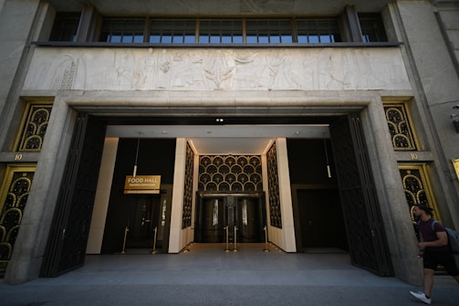 An ornate entranceway of a building is displayed, featuring art deco elements and golden accents. Above the entrance, there is a detailed relief sculpture, and a sign reads 'Food Hall Galeria Canalejas.' The doors are large and appear to be open, with decorative grids. A person walks by the front of the building, carrying a backpack.