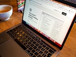 A laptop is open on a wooden table with a webpage displaying '100 ChatGPT audience building prompts' visible on the screen. In the background, there is a bowl of coffee, which seems partially full.