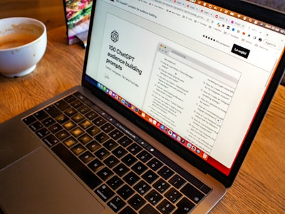 A laptop is open on a wooden table with a webpage displaying '100 ChatGPT audience building prompts' visible on the screen. In the background, there is a bowl of coffee, which seems partially full.