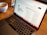 A laptop is open on a wooden table with a webpage displaying '100 ChatGPT audience building prompts' visible on the screen. In the background, there is a bowl of coffee, which seems partially full.