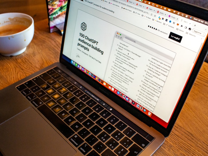 A laptop is open on a wooden table with a webpage displaying '100 ChatGPT audience building prompts' visible on the screen. In the background, there is a bowl of coffee, which seems partially full.
