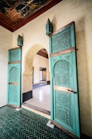 An ornate interior featuring intricately carved wooden doors painted in teal, adorned with geometric and floral patterns. The doors are set in an arched doorway with light-colored stone walls, and they lead into a room with a polished white floor. The ceiling above is decorated with an intricate, dark wood design. The floor in the foreground consists of green herringbone tiles.
