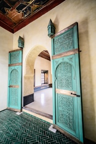 An ornate interior featuring intricately carved wooden doors painted in teal, adorned with geometric and floral patterns. The doors are set in an arched doorway with light-colored stone walls, and they lead into a room with a polished white floor. The ceiling above is decorated with an intricate, dark wood design. The floor in the foreground consists of green herringbone tiles.