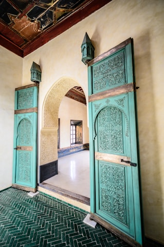 An ornate interior featuring intricately carved wooden doors painted in teal, adorned with geometric and floral patterns. The doors are set in an arched doorway with light-colored stone walls, and they lead into a room with a polished white floor. The ceiling above is decorated with an intricate, dark wood design. The floor in the foreground consists of green herringbone tiles.
