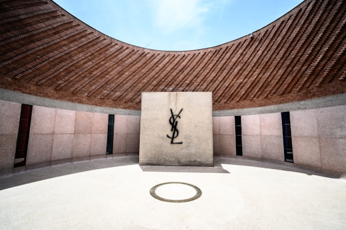 A circular architectural space with a curved brick roof and light-colored stone walls. At the center, a prominent logo is displayed on a standalone stone slab. The flooring is smooth and light-colored, with a circular design feature in front of the slab.