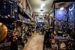 An intricately decorated shop filled with a variety of colorful items, including jewelry, vases, pottery, and decorative plates. The walls are adorned with hanging necklaces and trinkets against a vivid blue backdrop. The shelves are densely packed with decorative artifacts and handicrafts, creating a vibrant and cluttered atmosphere.