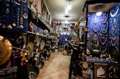 An intricately decorated shop filled with a variety of colorful items, including jewelry, vases, pottery, and decorative plates. The walls are adorned with hanging necklaces and trinkets against a vivid blue backdrop. The shelves are densely packed with decorative artifacts and handicrafts, creating a vibrant and cluttered atmosphere.