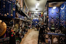 An intricately decorated shop filled with a variety of colorful items, including jewelry, vases, pottery, and decorative plates. The walls are adorned with hanging necklaces and trinkets against a vivid blue backdrop. The shelves are densely packed with decorative artifacts and handicrafts, creating a vibrant and cluttered atmosphere.