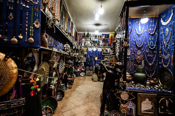 An intricately decorated shop filled with a variety of colorful items, including jewelry, vases, pottery, and decorative plates. The walls are adorned with hanging necklaces and trinkets against a vivid blue backdrop. The shelves are densely packed with decorative artifacts and handicrafts, creating a vibrant and cluttered atmosphere.