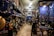 An intricately decorated shop filled with a variety of colorful items, including jewelry, vases, pottery, and decorative plates. The walls are adorned with hanging necklaces and trinkets against a vivid blue backdrop. The shelves are densely packed with decorative artifacts and handicrafts, creating a vibrant and cluttered atmosphere.