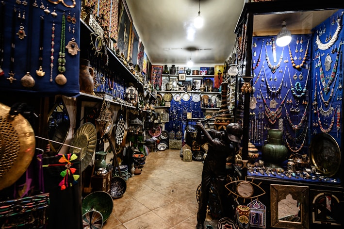 An intricately decorated shop filled with a variety of colorful items, including jewelry, vases, pottery, and decorative plates. The walls are adorned with hanging necklaces and trinkets against a vivid blue backdrop. The shelves are densely packed with decorative artifacts and handicrafts, creating a vibrant and cluttered atmosphere.