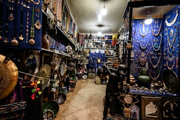 An intricately decorated shop filled with a variety of colorful items, including jewelry, vases, pottery, and decorative plates. The walls are adorned with hanging necklaces and trinkets against a vivid blue backdrop. The shelves are densely packed with decorative artifacts and handicrafts, creating a vibrant and cluttered atmosphere.