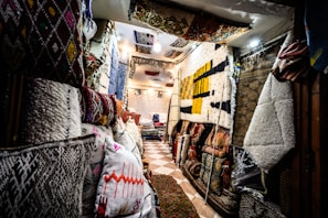 A narrow interior space filled with an array of colorful and patterned rugs and textiles displayed on the walls and on racks. The light is warm and creates a cozy atmosphere, with a tiled floor enhancing the ethnic and traditional vibe of the setting. Pillows and folded rugs are stacked neatly, showcasing intricate designs and vibrant colors.