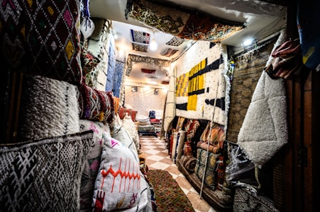 A narrow interior space filled with an array of colorful and patterned rugs and textiles displayed on the walls and on racks. The light is warm and creates a cozy atmosphere, with a tiled floor enhancing the ethnic and traditional vibe of the setting. Pillows and folded rugs are stacked neatly, showcasing intricate designs and vibrant colors.